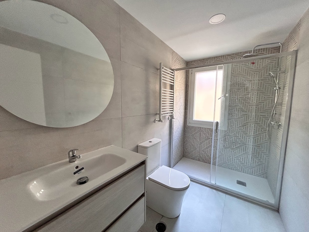 Resultado de reforma de baño en Madrid, con cerámicas rectificadas de gran tamaño, lavabo y espejo redondo, Wc con tapa amortiguadora y una amplia ducha con mampara corredera, junto a un toallero calefactor.
